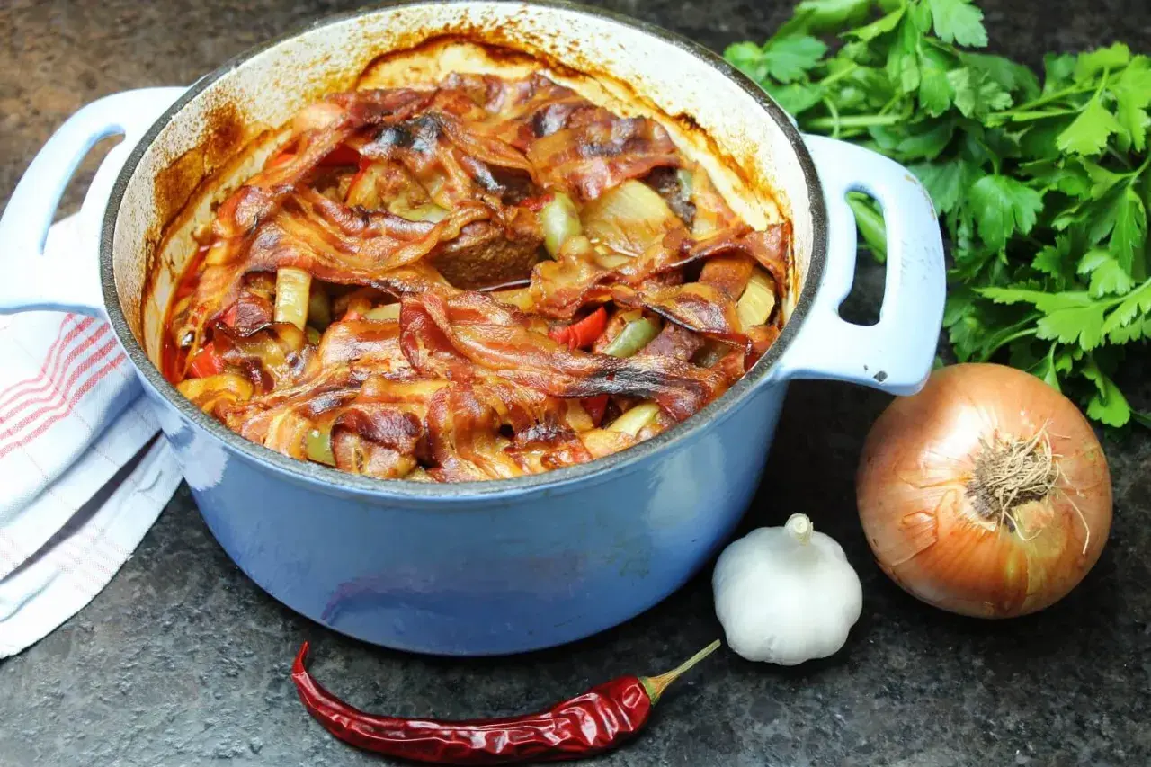 Herzhaftes Schichtfleisch im Backofen, bedeckt mit knusprigem Speck, daneben Zwiebel, Knoblauch und Chili.