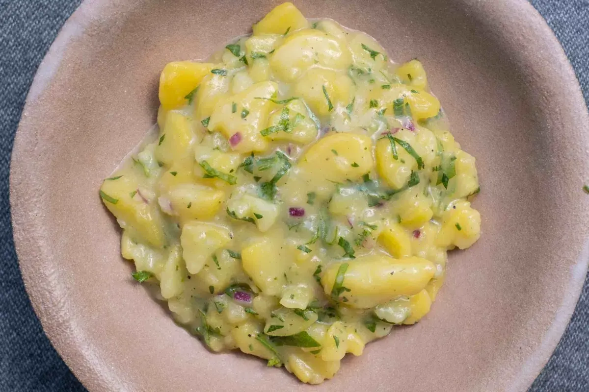 Ein cremiger Kartoffelsalat mit Speck, verfeinert mit frischer Petersilie und roten Zwiebeln, serviert in einer braunen Schale.