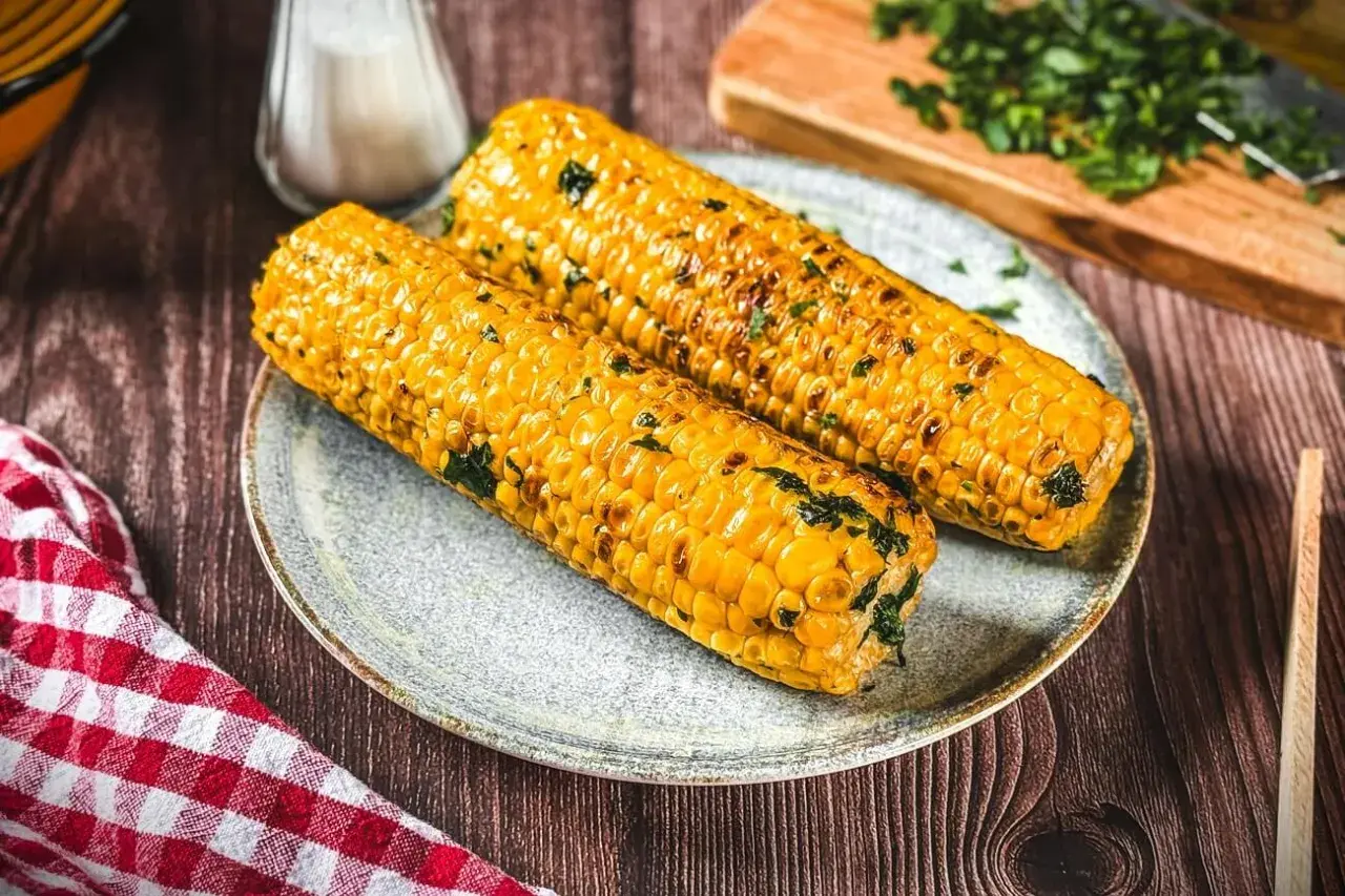Zwei goldgelbe Maiskolben, perfekt gegrillt und mit Kräutern bestreut, liegen auf einem Teller. Ein Genuss für alle Sinne!