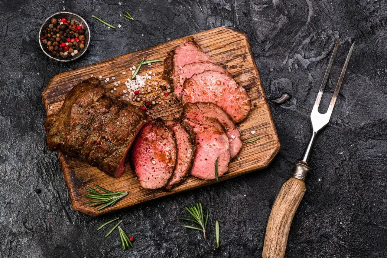 Saftiges Roastbeef, perfekt gegrillt und in Scheiben geschnitten, liegt auf einem Holzbrett. Gewürze und Rosmarin garnieren das Gericht.