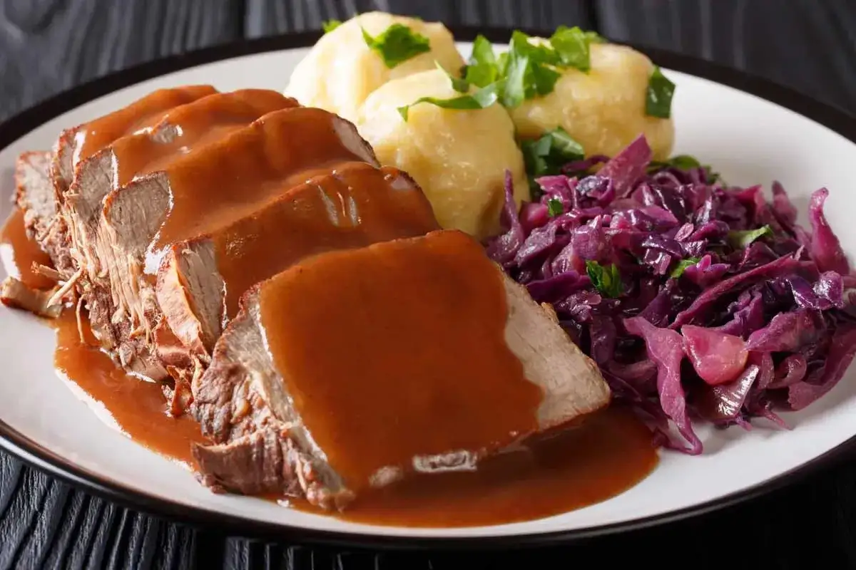 Ein Teller mit Scheiben von saftigem Sauerbraten, übergossen mit dunkler Soße, dazu Klöße und Rotkohl. Ein klassisches Sauerbraten Rezept.