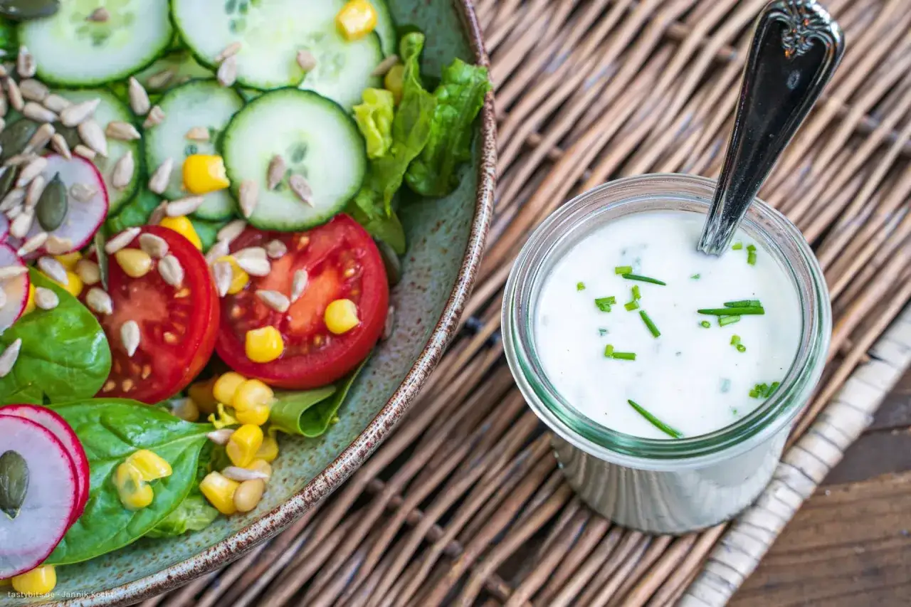 Frischer Salat mit Tomaten, Gurken und Mais, daneben ein Glas mit selbstgemachtem Joghurt-Dressing, garniert mit Schnittlauch.