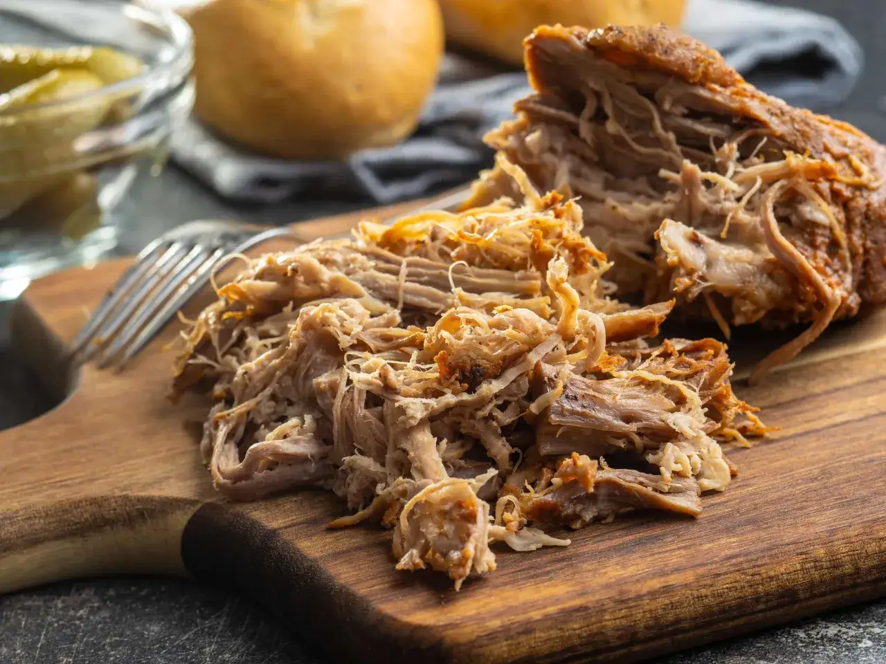 Zartes pulled pork sous vide auf einem Holzbrett, bereit für Burger oder Sandwiches. Daneben liegen Brötchen und Gurken.