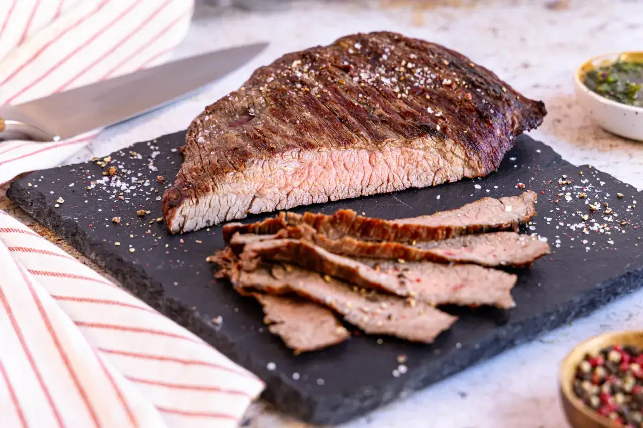 Perfekt gegrilltes Flanksteak, auf einem Schieferbrett serviert, mit grobem Salz und Pfeffer bestreut. Die Scheiben zeigen einen zarten rosa Kern.