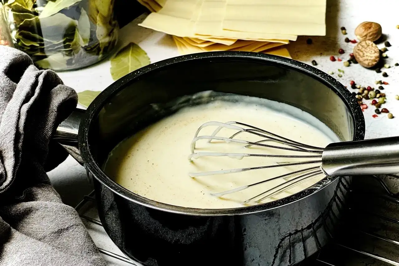 Cremige B&eacute;chamelsauce in einem Topf, bereit f&uuml;r Lasagne. Daneben liegen Lasagneplatten, Lorbeerbl&auml;tter und Gew&uuml;rze.