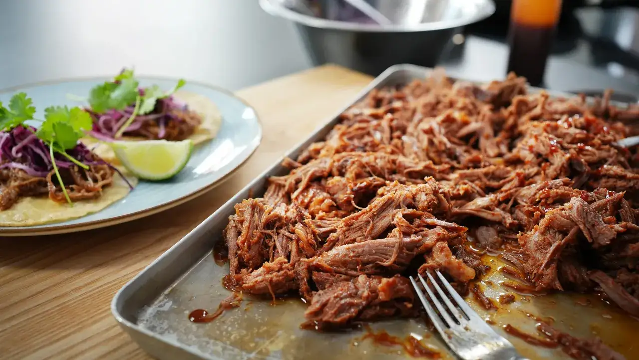Saftiges Pulled Pork, perfekt für ein Smoker-Rezept, liegt auf einem Blech. Daneben zwei Tacos mit dem Fleisch, Rotkohl und Koriander.