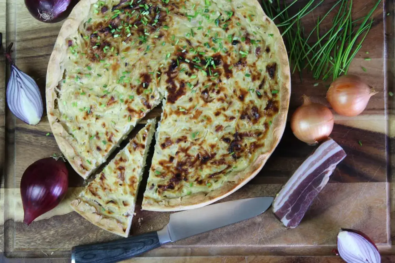 Ein goldbrauner Zwiebelkuchen, frisch aus dem Ofen, mit Schnittlauch bestreut. Zutaten f&uuml;r das Zwiebelkuchen Rezept liegen daneben.