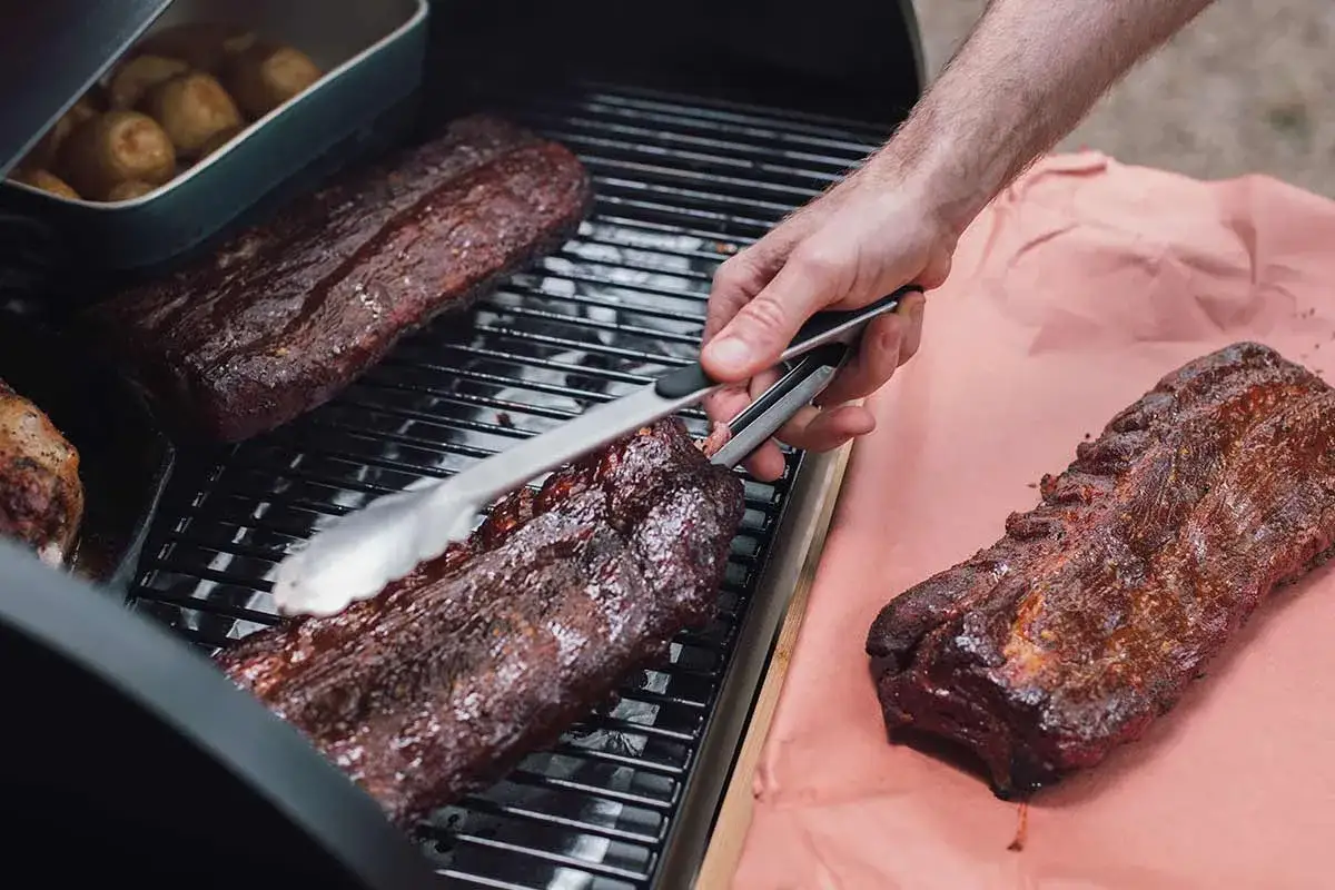 Spareribs werden auf dem Grill zubereitet. Ein St&uuml;ck liegt auf Butcher Paper, ein anderes wird mit einer Zange gewendet.