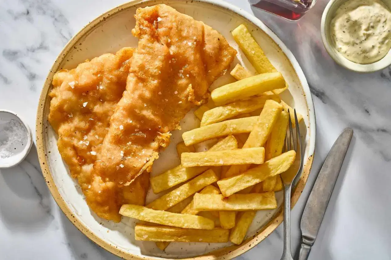 Knuspriges Fish and Chips auf einem Teller, bestreut mit grobem Salz. Daneben eine Sch&uuml;ssel mit Tartarensauce und ein Glas Malzessig.