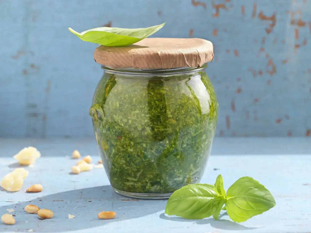 Glas mit selbstgemachtem Basilikum Pesto, garniert mit Basilikumblatt. Pinienkerne und Parmesan liegen daneben.