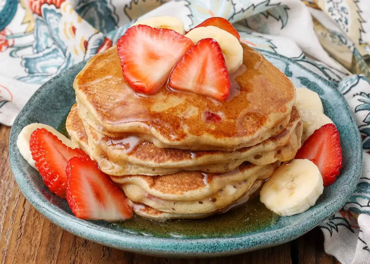 Stapel fluffiger Bananen Pancakes ohne Ei, garniert mit frischen Erdbeeren und Bananenscheiben, betr&auml;ufelt mit Sirup.