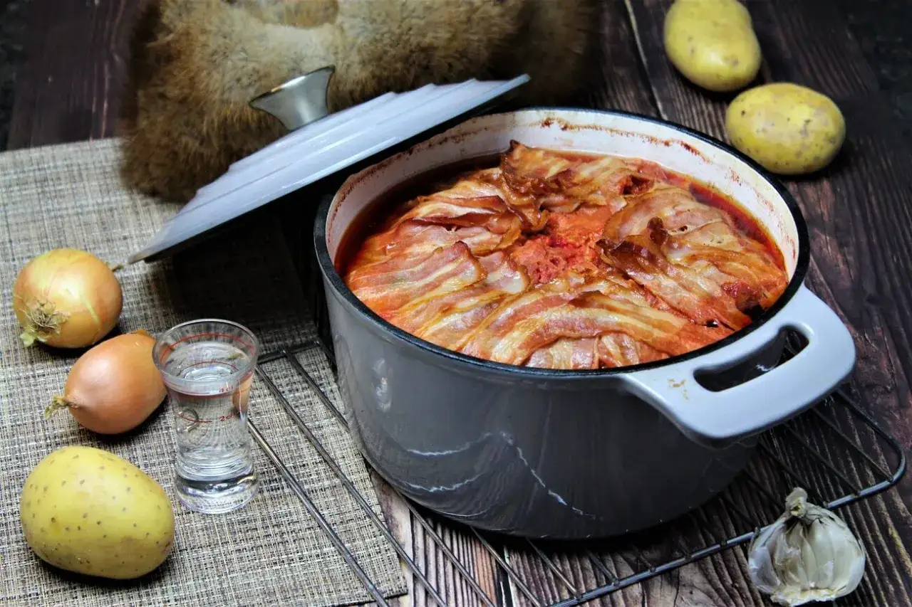 Herzhaftes Gericht in einem Topf, bedeckt mit Speckstreifen. Dazu Zwiebeln, Kartoffeln und ein Schnapsglas – typisch für russische Gerichte.