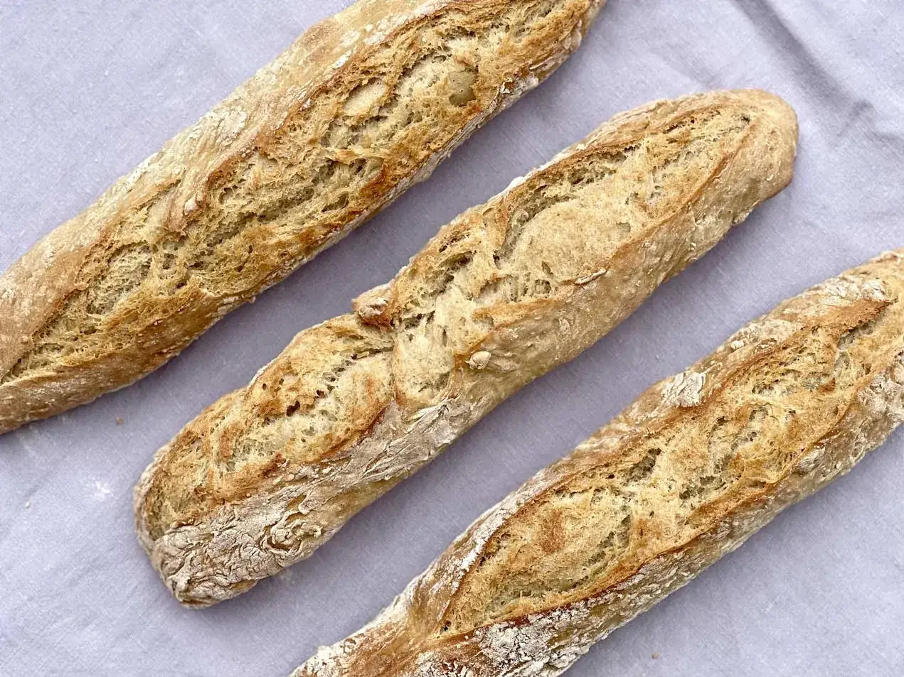 Drei frisch gebackene Baguettes liegen nebeneinander auf einem grauen Tuch. Das knusprige Baguette-Rezept ist perfekt gelungen.