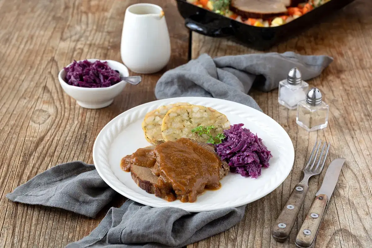 Saftiger rinderbraten im Dutch Oven, serviert mit Kl&ouml;&szlig;en und Rotkohl auf einem rustikalen Holztisch.
