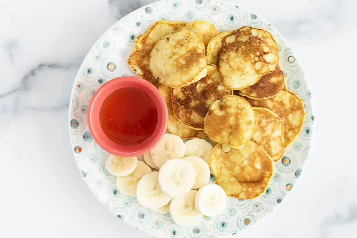 Kleine, goldbraune Bananen-Pancakes mit Bananenscheiben und Honig auf einem Teller. Perfekt f&uuml;r ein Baby-Fr&uuml;hst&uuml;ck.