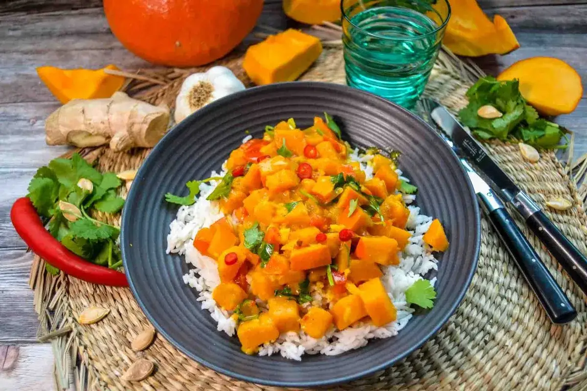 Herzhafte Hokkaido-Rezepte: Ein bunter Kürbis-Curry mit Reis, garniert mit Koriander und Chili. Dazu Ingwer, Knoblauch und ein Glas Wasser.