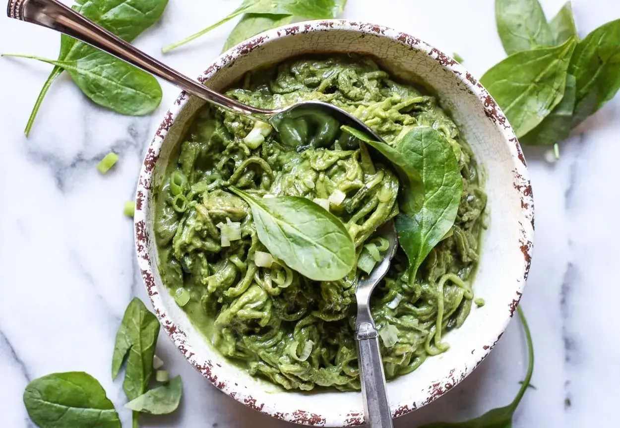 Sch&uuml;ssel mit cremiger, gr&uuml;ner Pasta, garniert mit frischem Spinat und Fr&uuml;hlingszwiebeln. Ein L&ouml;ffel ragt heraus, bereit, das k&ouml;stliche Avocado-Pesto zu genie&szlig;en.