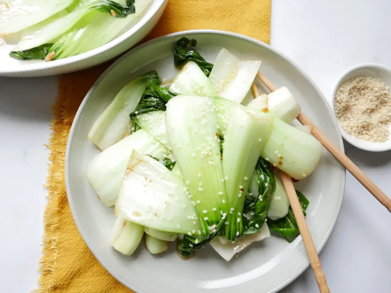 Ged&auml;mpfter Pak Choi mit Sesam bestreut, serviert mit St&auml;bchen auf einem Teller. Daneben eine Schale mit Sesam.