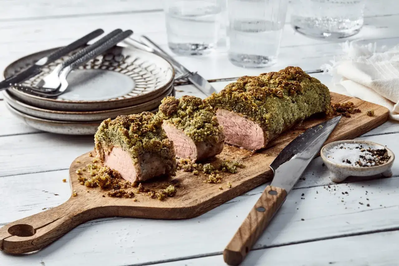 Saftiges schweinefilet rezept mit Kr&auml;uterkruste, auf Holzbrett serviert. Daneben Messer, Salz und Pfeffer.