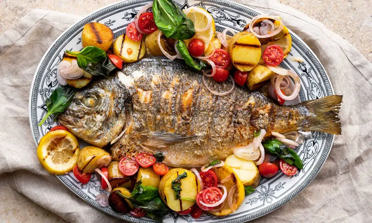 Ganze Dorade im Backofen, goldbraun gebraten, serviert mit gegrillten Kartoffeln, Kirschtomaten, Zitronenspalten und roten Zwiebelringen.
