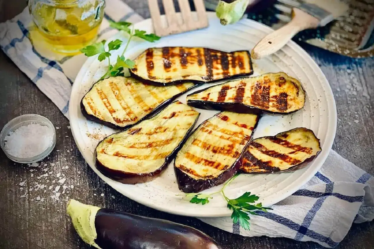 Gegrillte Auberginenscheiben auf einem Teller, bereit für köstliche Rezepte mit Auberginen. Daneben Salz, Öl und frische Petersilie.
