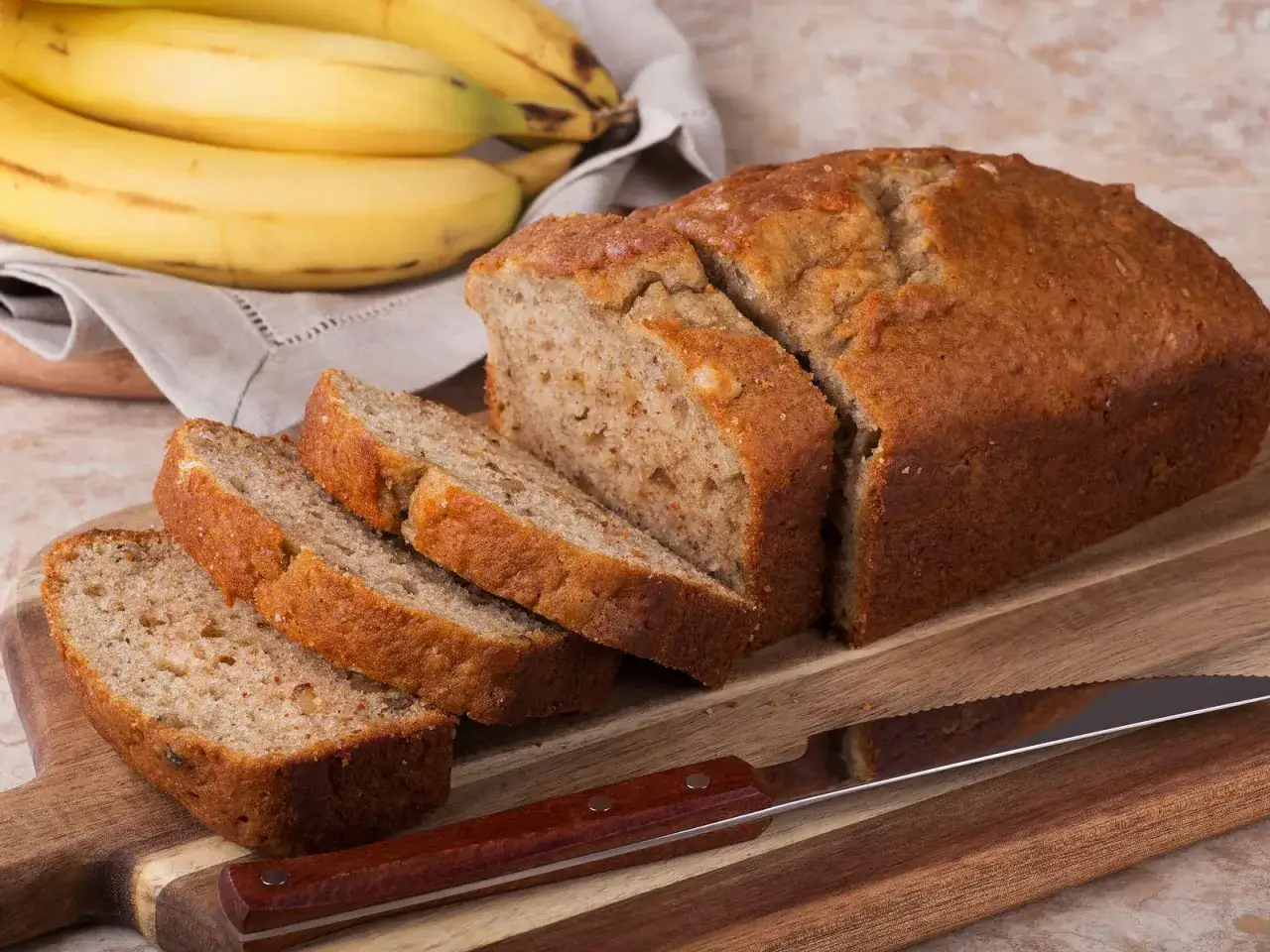 Ein saftiges Bananenbrot ohne Zucker, frisch aufgeschnitten auf einem Holzbrett. Daneben liegen reife Bananen auf einem Leinentuch.