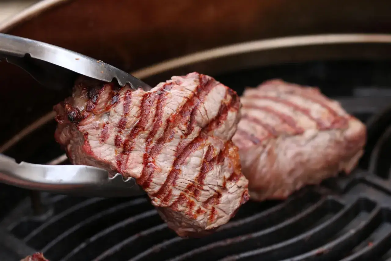Zangen halten ein saftiges Roastbeef mit Grillstreifen &uuml;ber einem Grillrost. Ein weiteres St&uuml;ck liegt daneben.