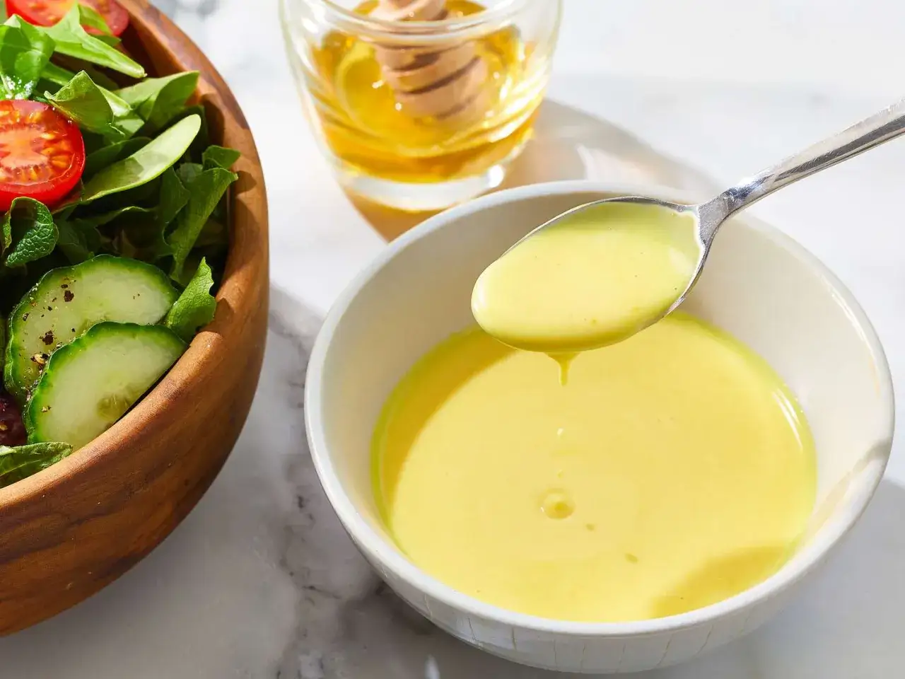 Ein L&ouml;ffel cremiges honig senf dressing tropft in eine Sch&uuml;ssel. Daneben ein Salat mit Tomaten und Gurken.