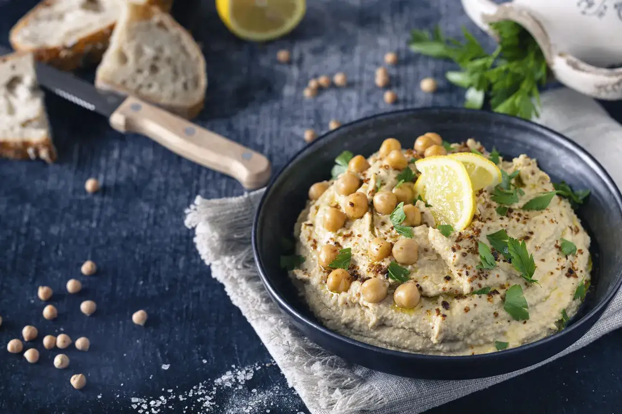 Cremiger Hummus, garniert mit Kichererbsen, Zitrone und Petersilie. Perfekt für ein einfaches Hummus-Rezept. Dazu gibt es Brot und ein Messer.